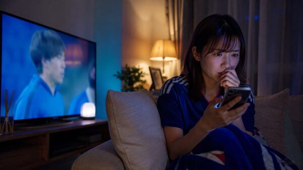 夜のリビングでサッカー中継を見ながら遠藤航の代表離脱と引退情報をスマホで確認する日本人女性